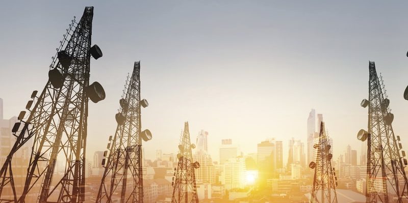 Torres de telecomunicaciones con antenas sobre una ciudad al atardecer.